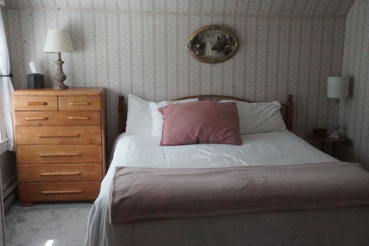 Ein Schlafzimmer mit einem hölzernen Schminktisch, einem Bett mit weißen Leinen und einem rosafarbenen Kissen, einem runden Spiegel an der Wand und einer Lampe auf dem Schminktisch.