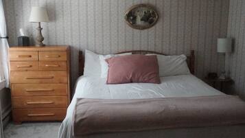 Um quarto com cômoda de madeira, cama com lençóis brancos e um travesseiro rosa, espelho redondo na parede e uma luminária sobre a cômoda.