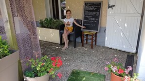 Iemand zit op een stoel voor een krijtbordmenu, met potplanten en een witte deur op de achtergrond.