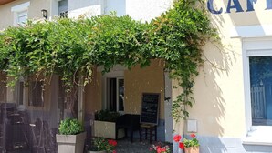 Een café met een pergola begroeid met groen, terrasstoelen en een krijtbordmenu.