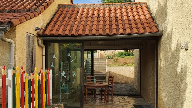 A patio area with a tiled roof, a glass door entrance, a wooden table and chairs, and a small garden with mulch and plants. - Maison Familiale, South Aveyron 5minutes from St Rome de Tarn beach  (Saint-Rome-de-Tarn)