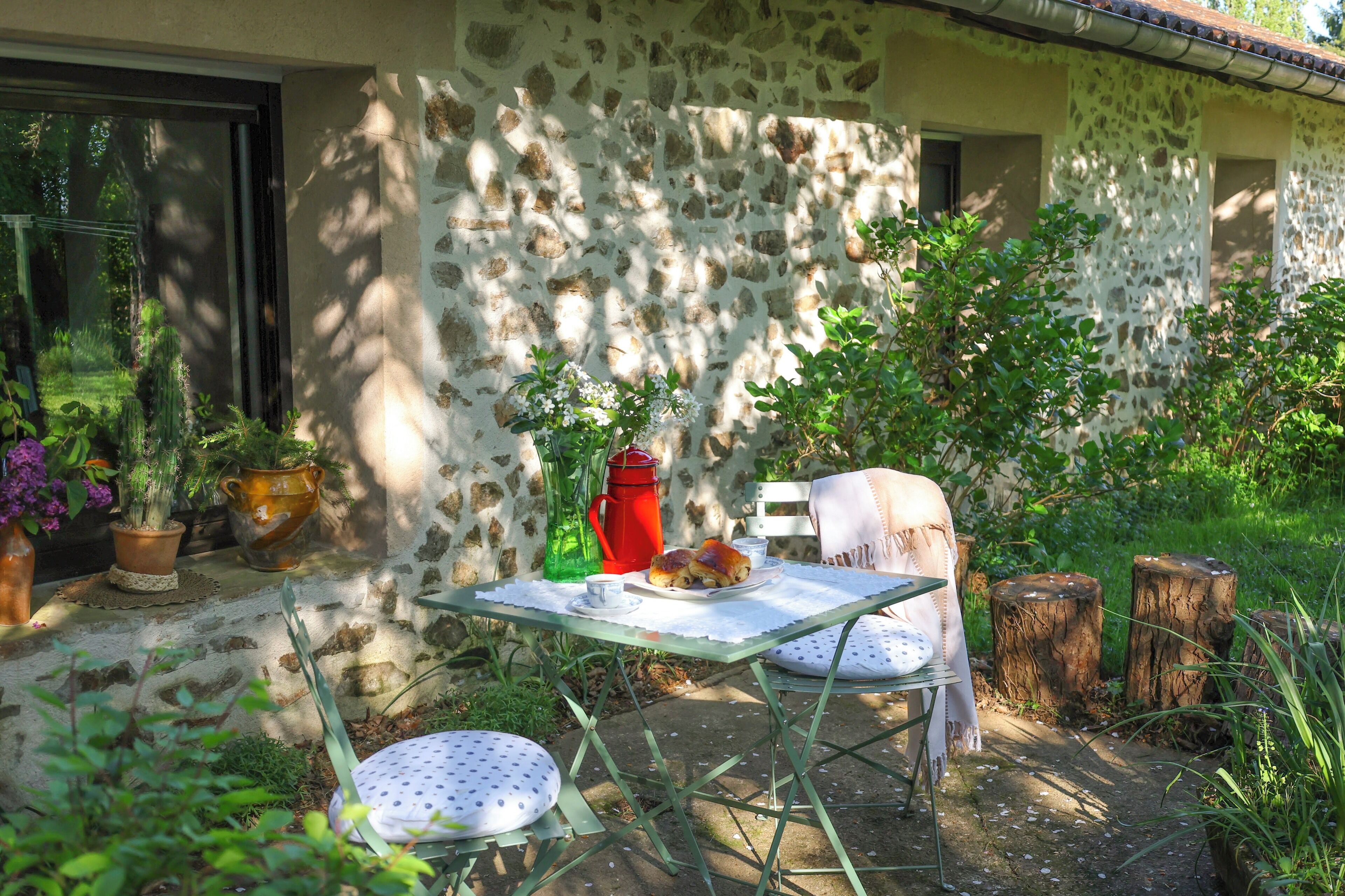 A stone building with a table and chairs outside, surrounded by greenery.