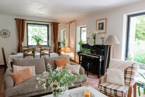 Ein Wohnzimmer mit einer Couch, Sesseln, einem Klavier und einem Essbereich mit Stühlen und einem Tisch.
