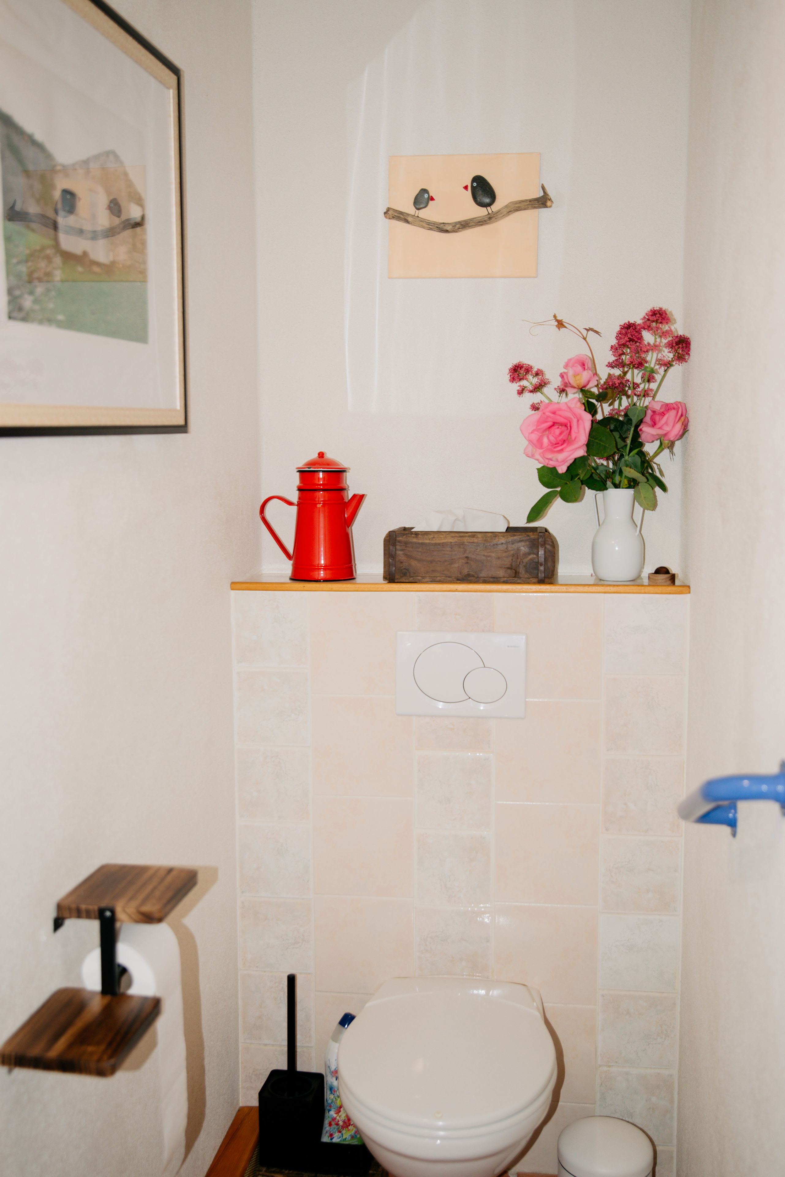 Un baño con inodoro, una tetera roja, un caja de madera, un florero con flores e interruptor de luz en la pared.