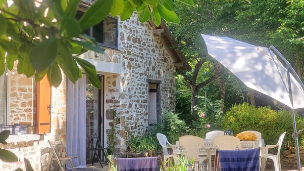 Un bâtiment en pierre doté d’un balcon, d’un espace extérieur aménagé avec des chaises et une table, et d’un grand parasol offrant de l’ombre.