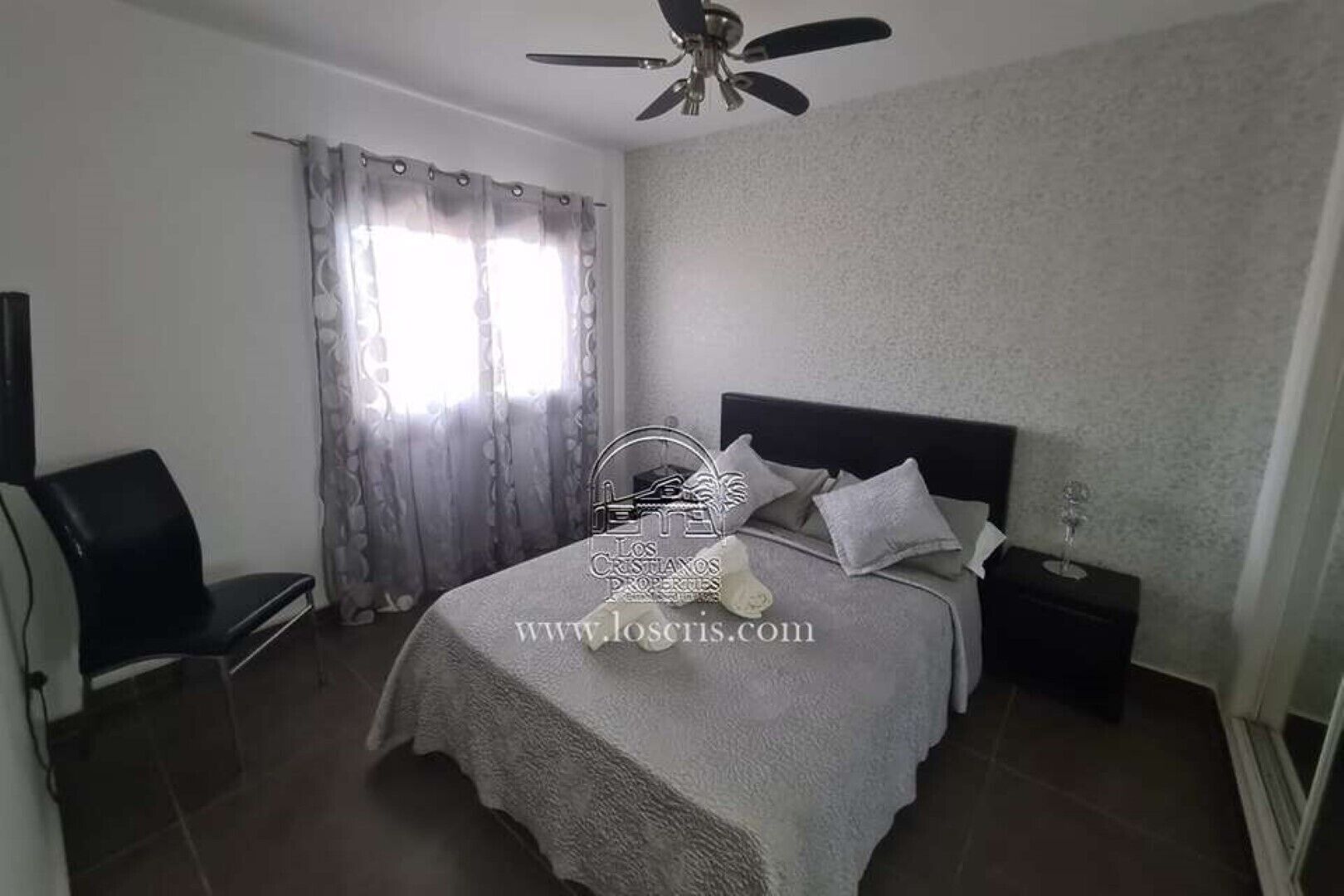 Une chambre à coucher comprenant un lit, une table de chevet, une chaise, un ventilateur de plafond et une fenêtre avec des rideaux.