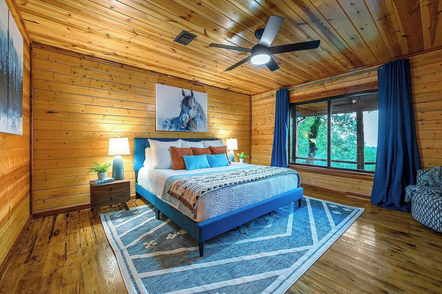 Un dormitorio con paneles de madera, una cama, dos mesitas de noche, un ventilador de techo y un amplio ventanal con cortinas.