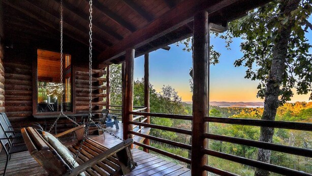 Um deck de madeira com uma cadeira de balanço, uma mesa e cadeiras, com vista para uma paisagem arborizada.