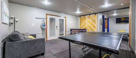 A room with a ping pong table, a couch, and a door with a mirror.