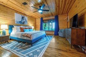 Un dormitorio de cabaña de madera con cama, ventilador de techo, televisor y ventana con cortinas.
