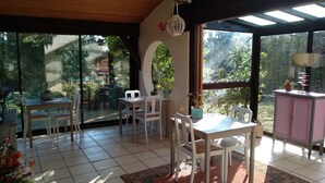 A sunlit patio with a dining table and chairs, surrounded by large windows offering a view of the garden. - Agréable Chambre Indépendante Avec Piscine & Parc (Brézilhac)