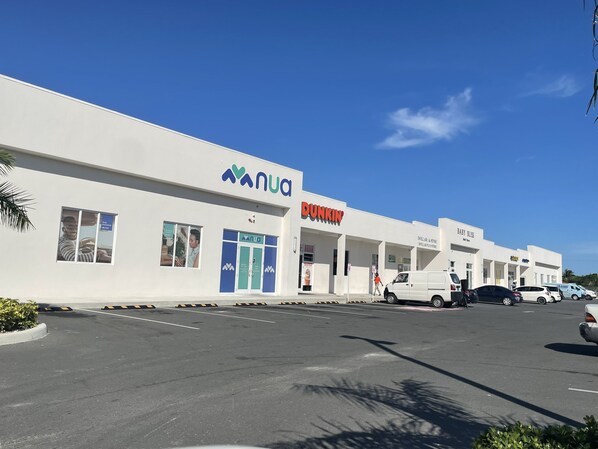Un edifici comercial amb els cartells 'MANUA' i 'DUNKIN', un pàrquing i un cel blau clar.