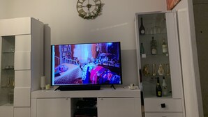 A living room with a flat-screen TV on a white entertainment unit, a clock on the wall, and a wine cabinet to the right. - Chambre Simple Privée -saint Priest (Saint-Priest)