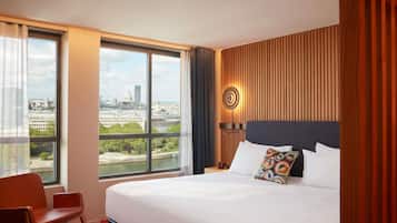 A hotel room with a large bed, a chair, a view of a cityscape, and a wooden panel wall.