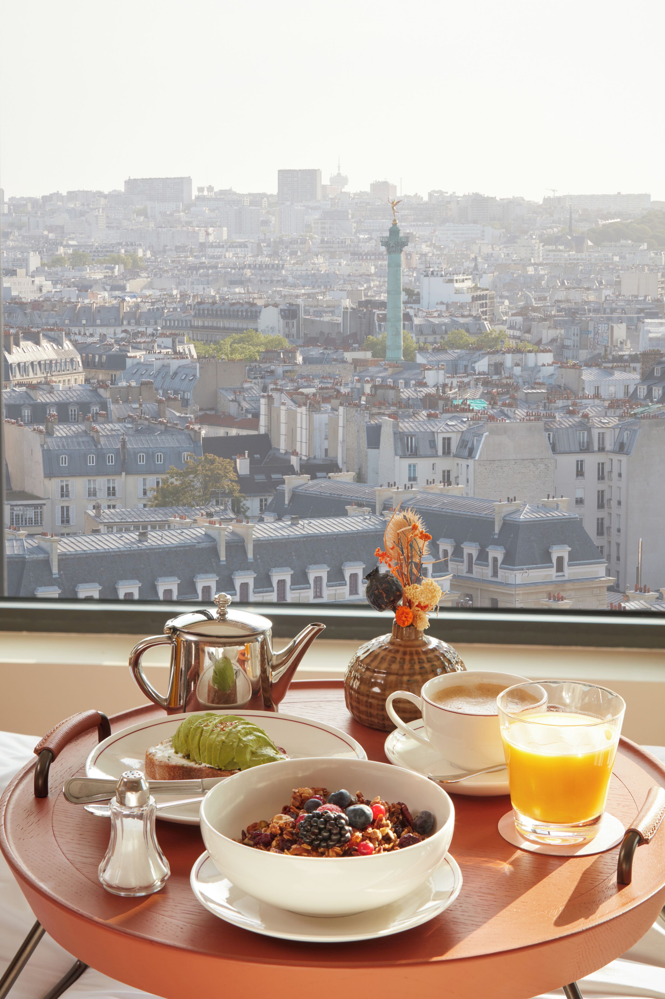 Bandeja de desayuno con un tazón de cereal, una taza de café y un vaso de jugo de naranja, colocada frente a una ventana con vista a un paisaje urbano.