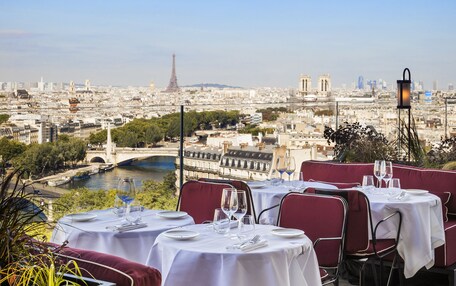 An outdoor dining area with tables set for two, overlooking a cityscape with the Eiffel Tower in the distance.