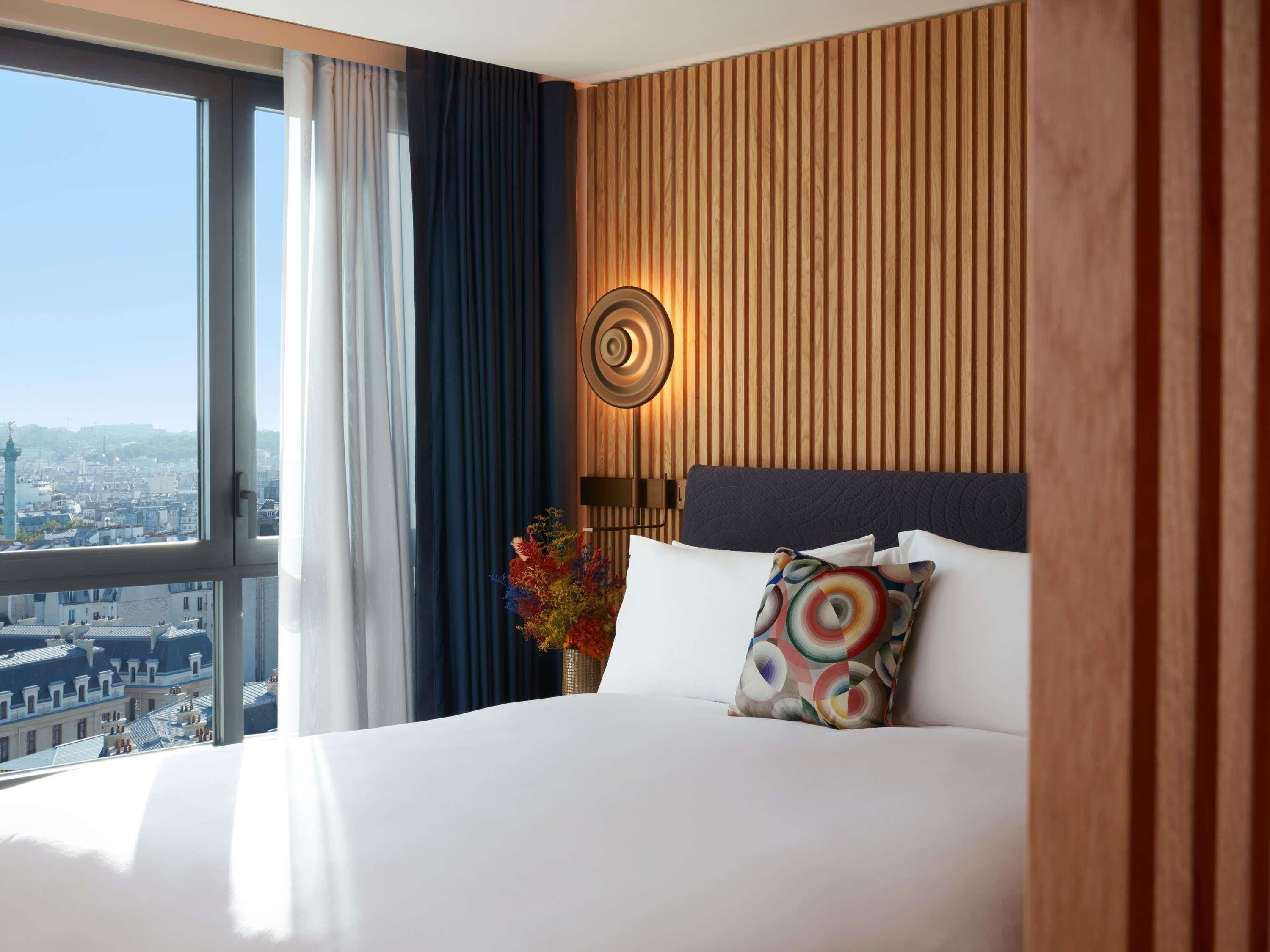 A hotel room with a large window, a bed with white linens, a wooden panel wall, and a decorative wall lamp.
