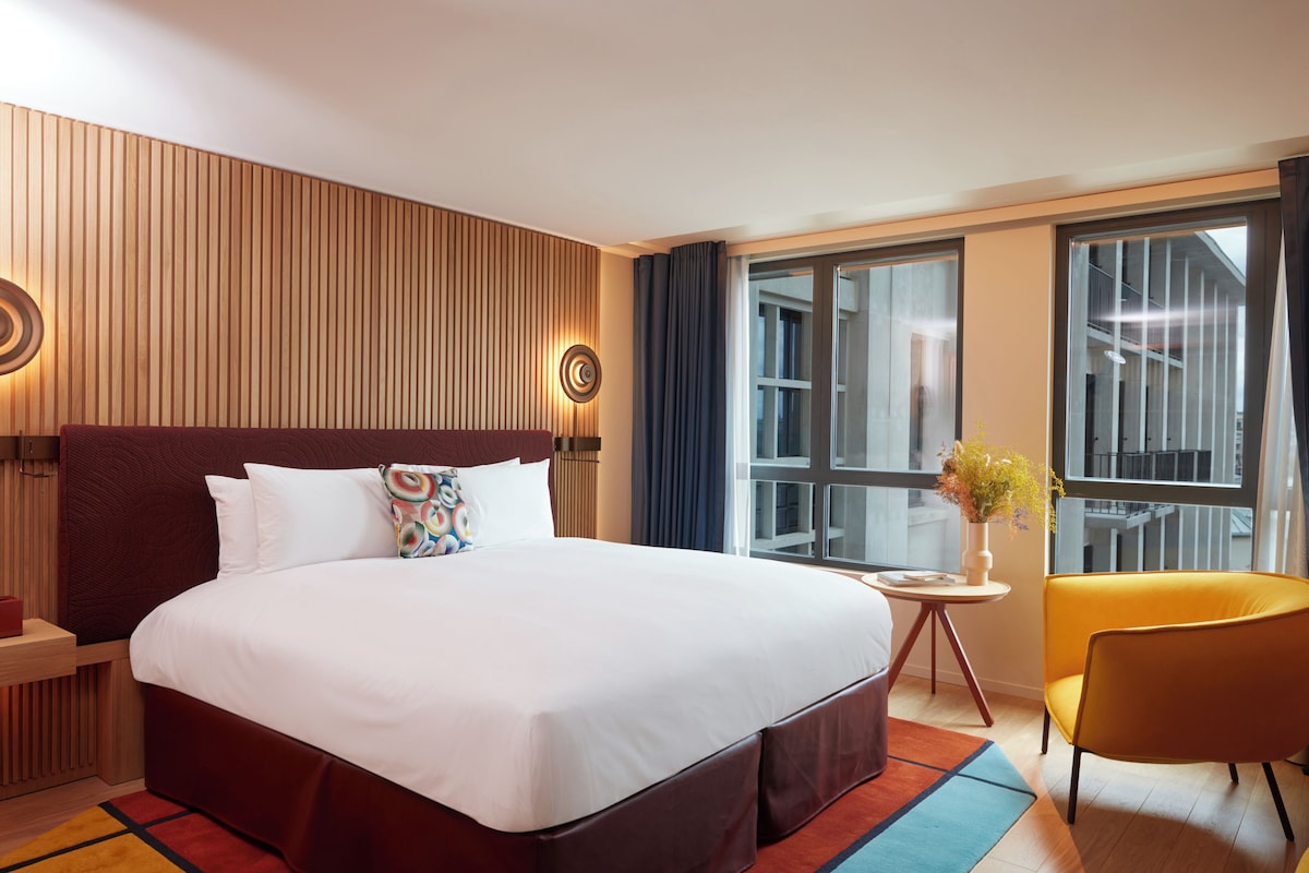 A hotel room with a large bed, a small table, a chair, and a window with a view.