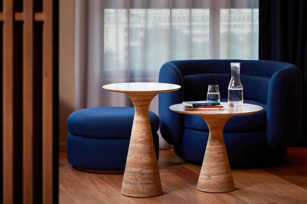 A modern room with two wooden tables, a blue armchair, and a window with a view of buildings.