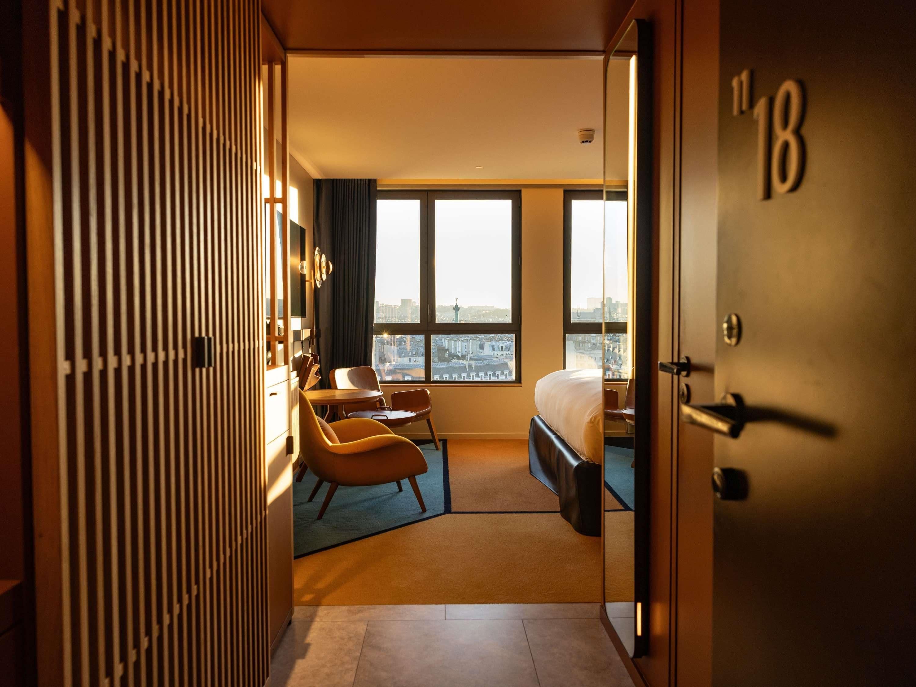 A hotel room with a bed, a desk, a chair, a wardrobe, and a view of the city.