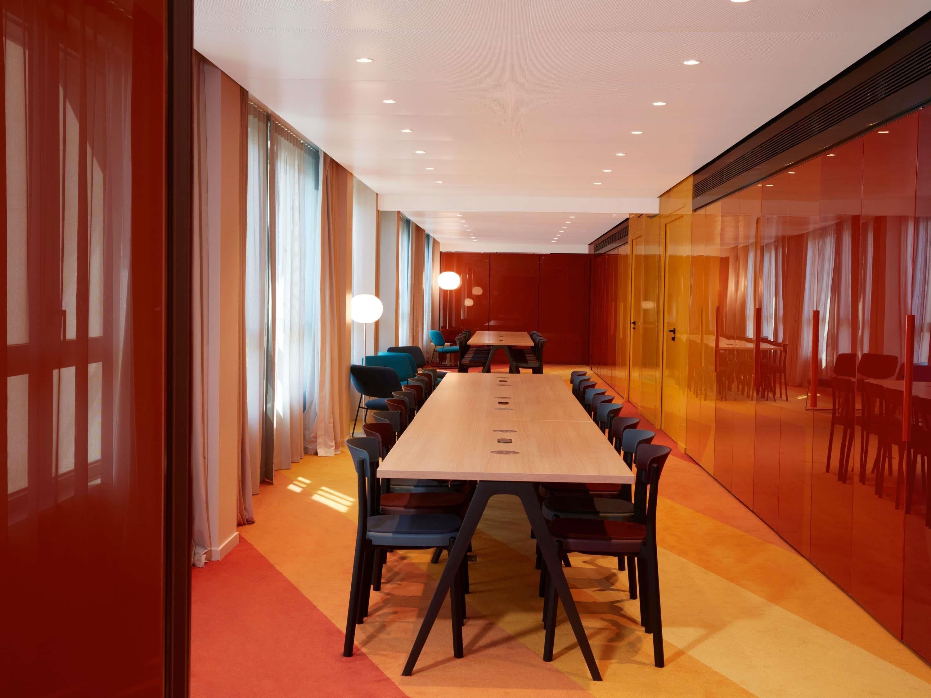 Uma sala de conferências com uma mesa comprida e cadeiras, janelas amplas com cortinas e um chão coberto por carpete vermelho.