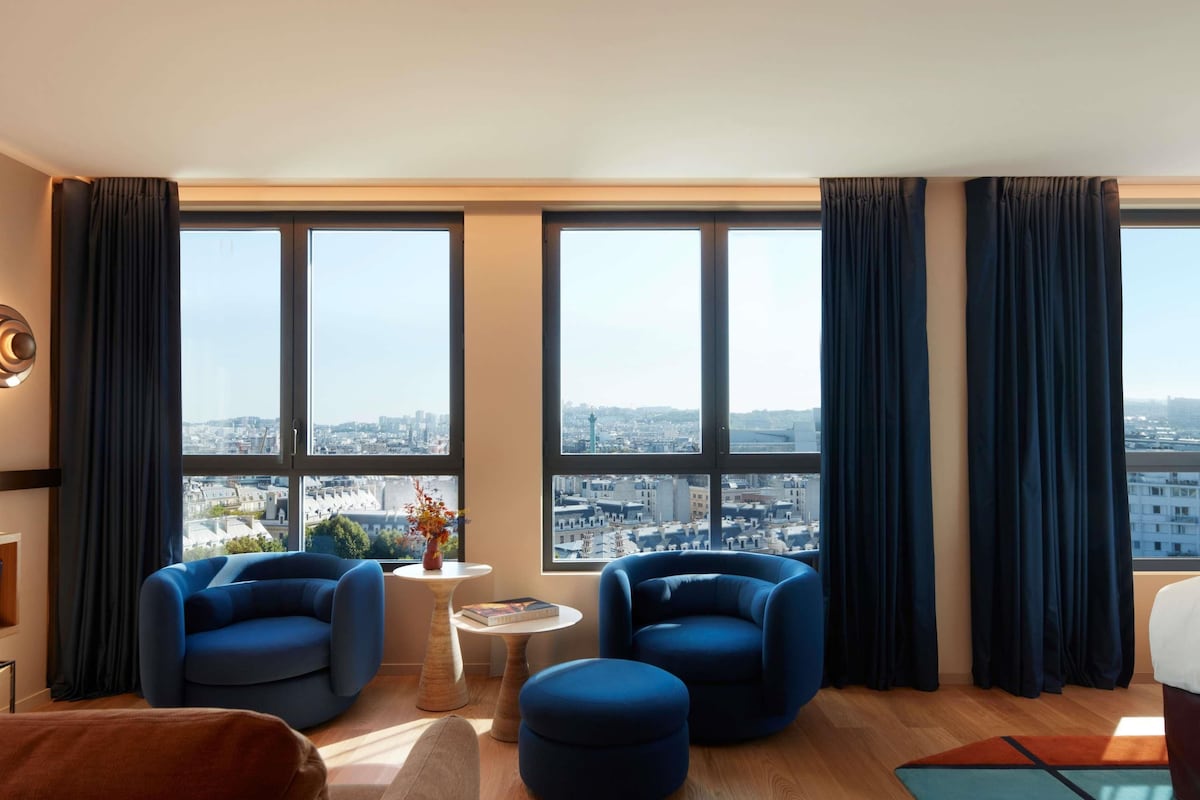 Ein modernes Wohnzimmer mit Stadtblick, ausgestattet mit zwei blauen Sesseln, einem kleinen runden Tisch mit einem Buch und einem braunen Sofa.
