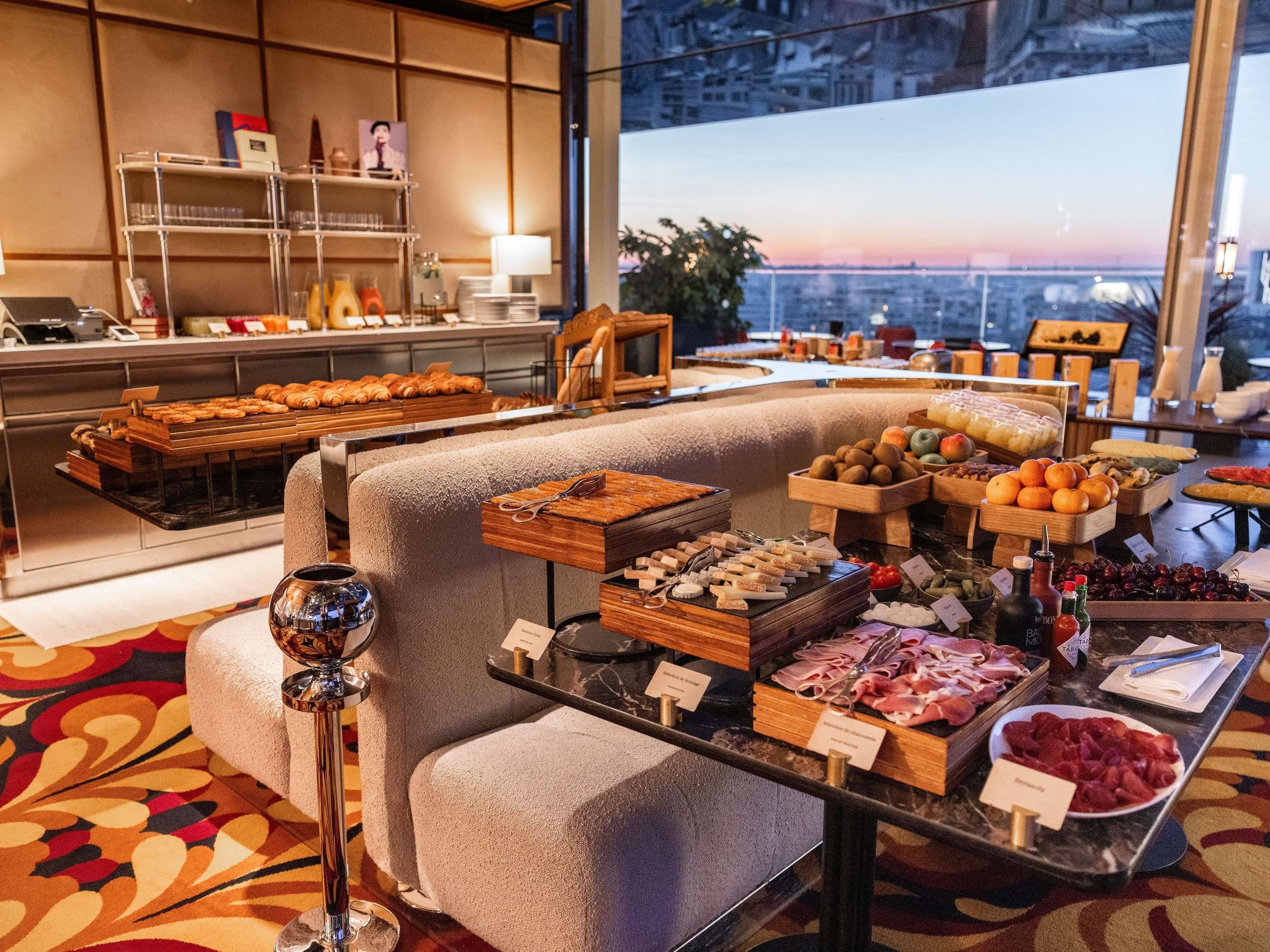 Buffet com diversos alimentos, um espaço para café e vista panorâmica da cidade através de amplas janelas.