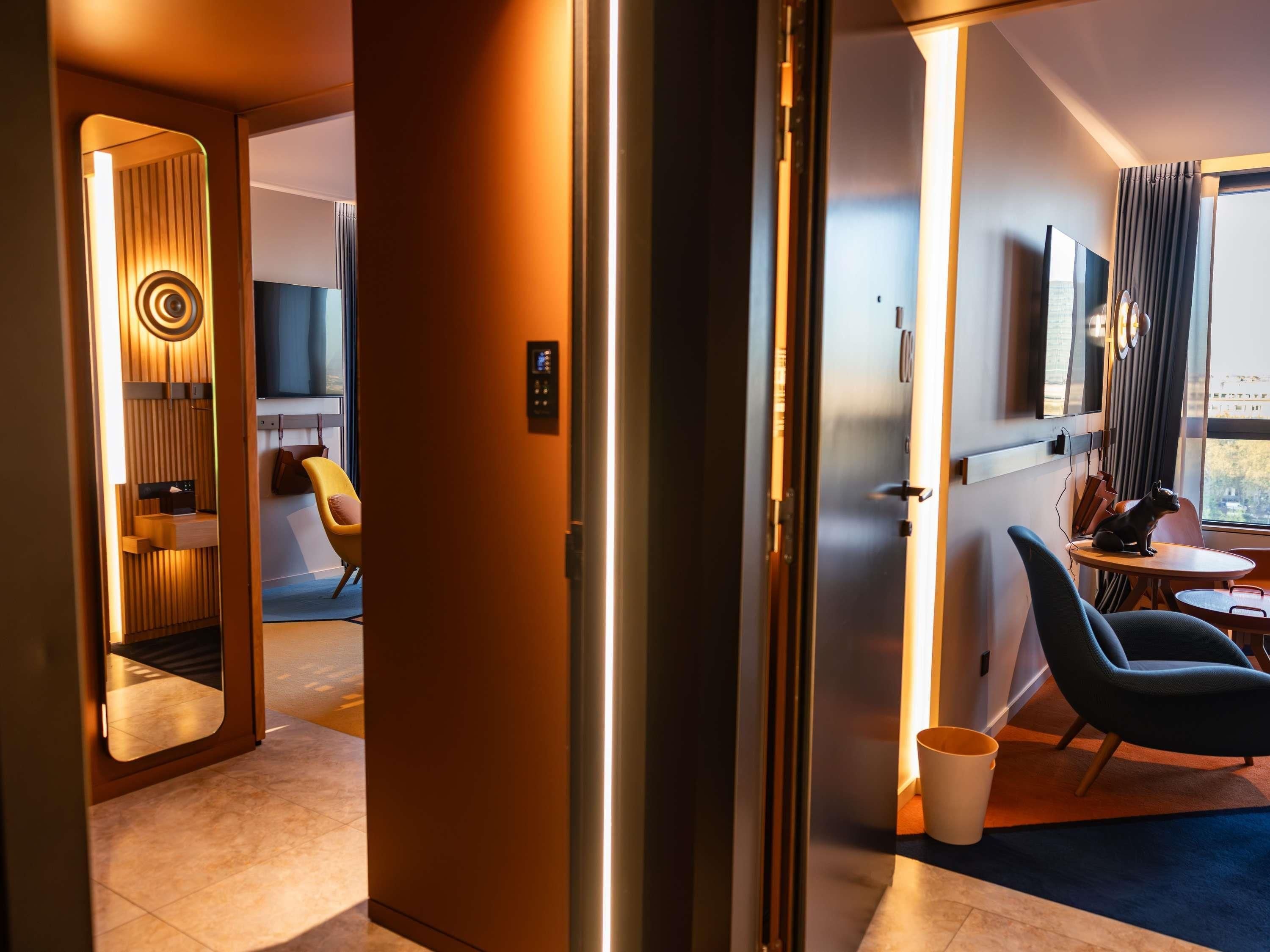 A modern hotel room with a large mirror, a television, a sofa, and a dining area.