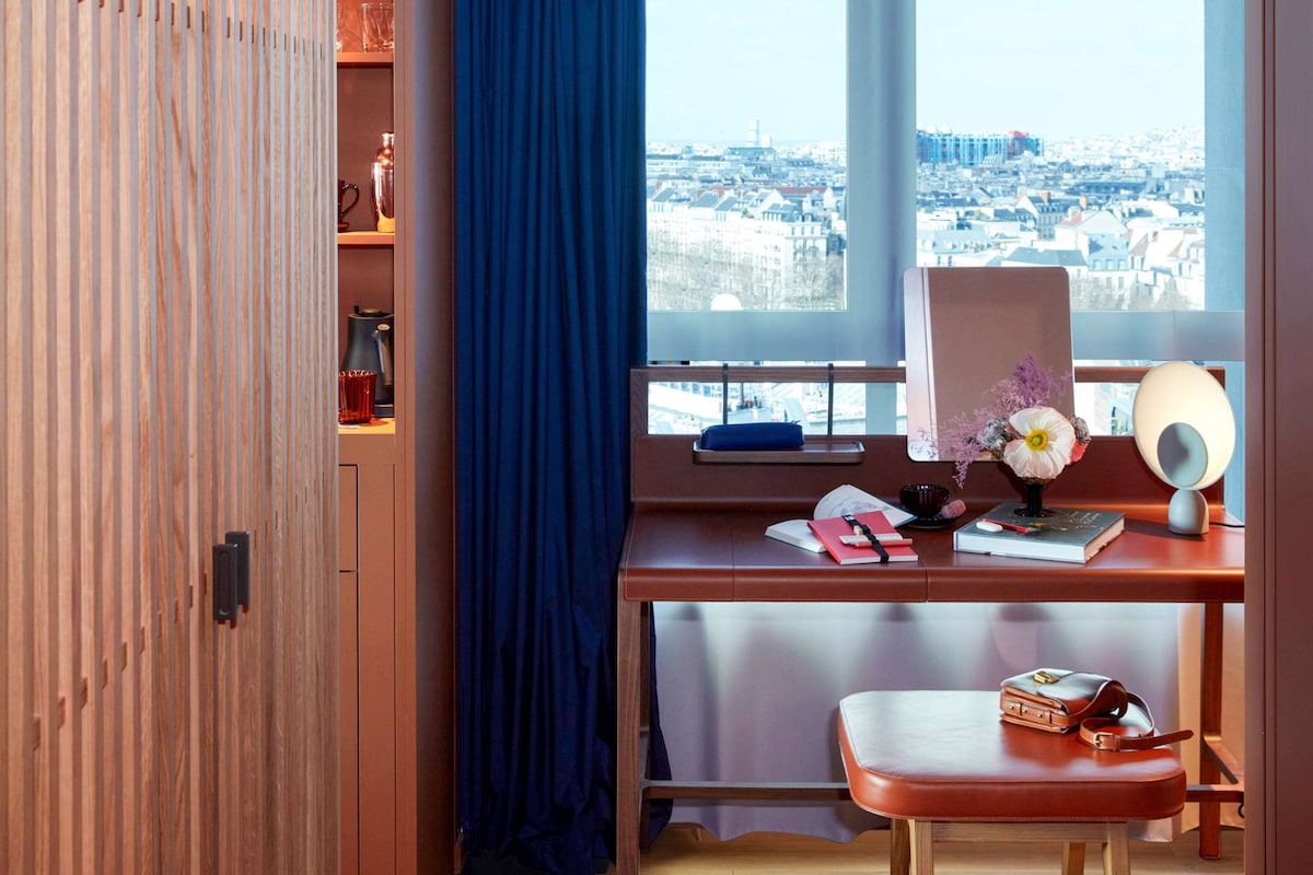 A room with a desk, a window with a city view, a blue curtain, and a wooden wardrobe.