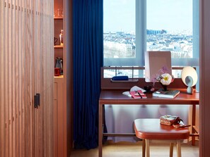 A room with a desk, a window with a city view, a blue curtain, and a wooden wardrobe.