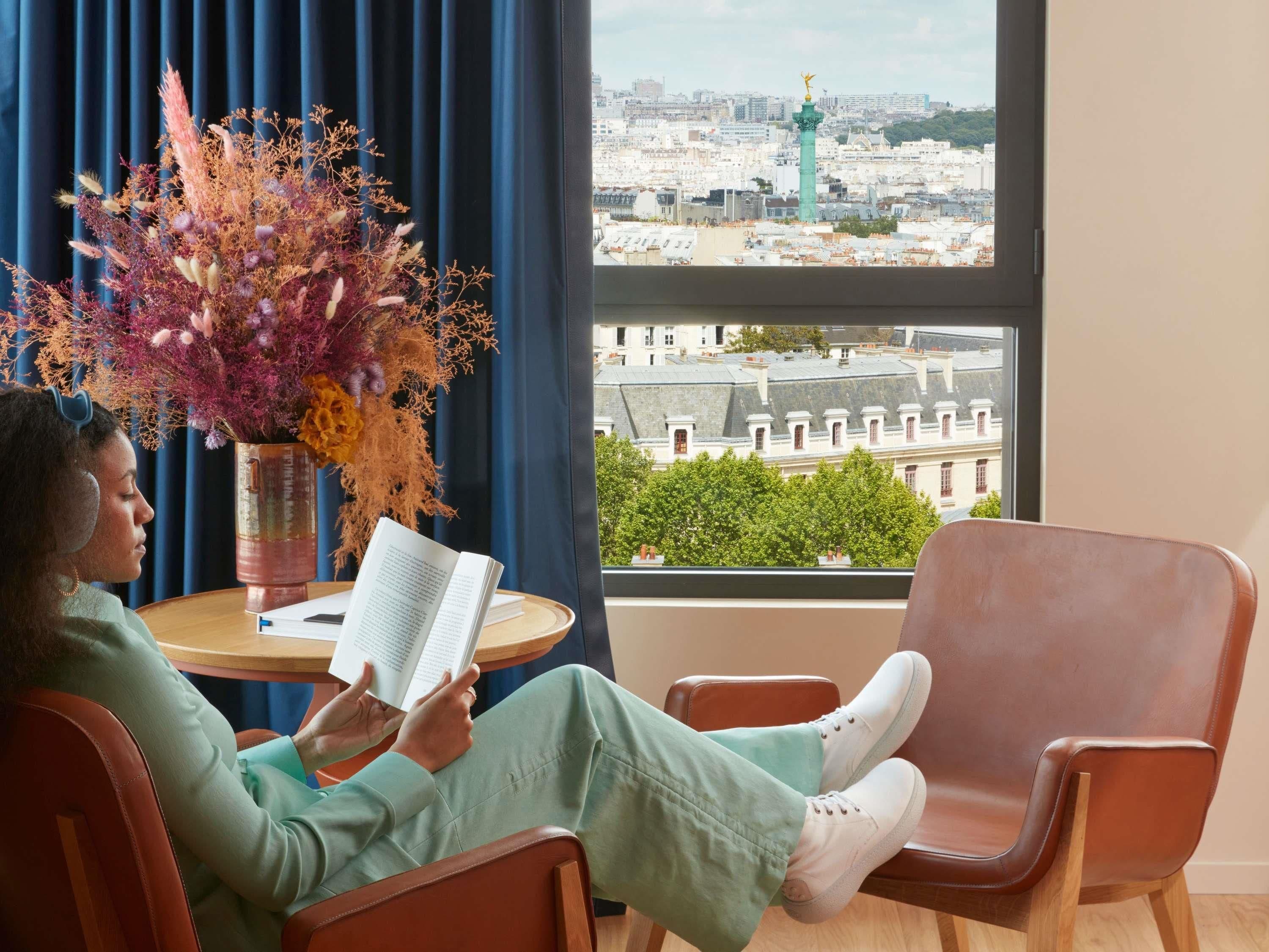 A person reading a book in a room with a view of a cityscape and a statue.