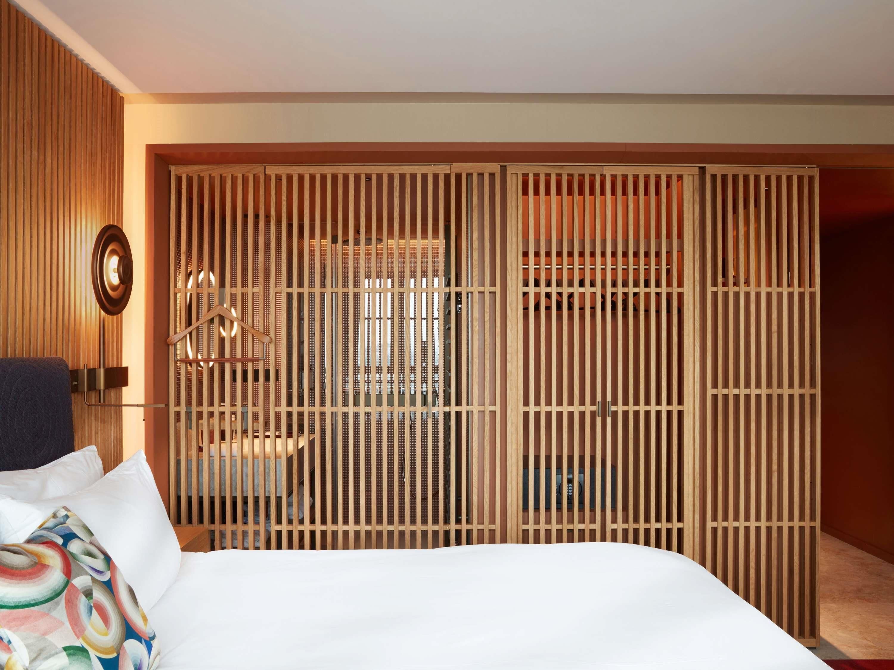 Una habitación de hotel con una cama, un separador de madera con puerta corredera, una lámpara fijada a la pared y una pequeña mesa con una silla.