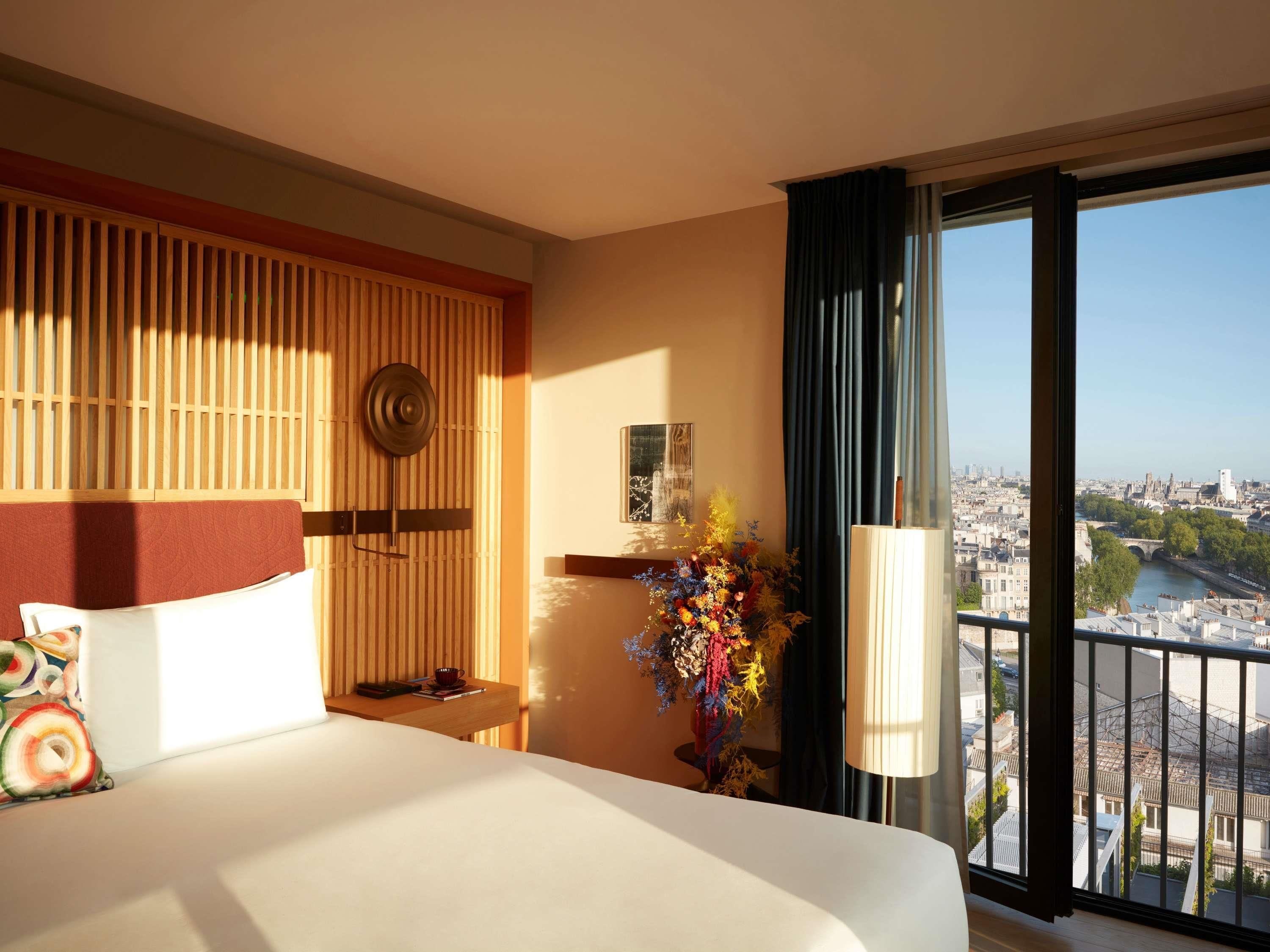 A hotel room with a bed, a wooden headboard, a decorative wall panel, a window with a view of a cityscape, and a vase with flowers.