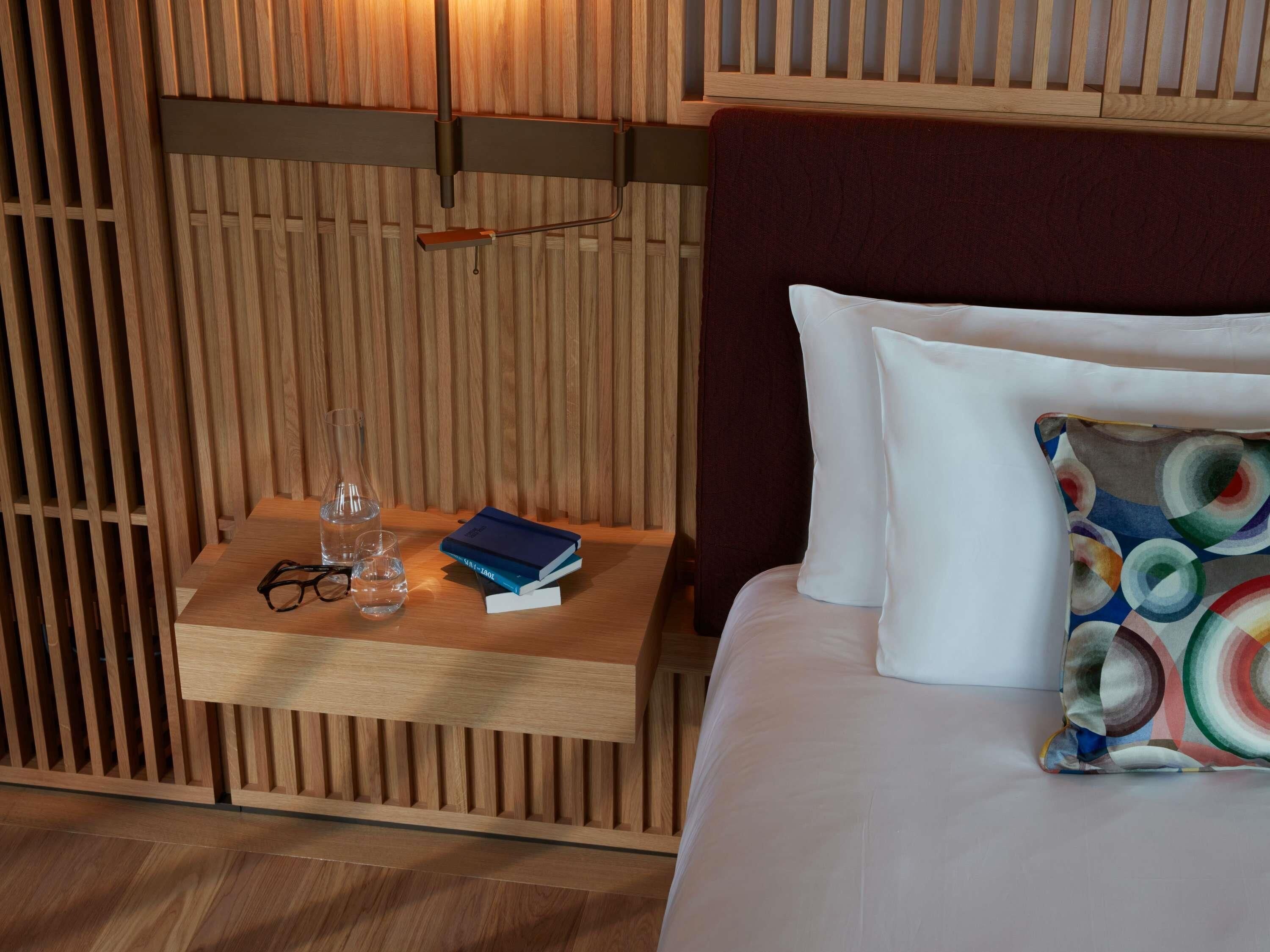 A neatly arranged hotel room with a wooden nightstand, a bed with white and colorful pillows, and a wooden headboard.