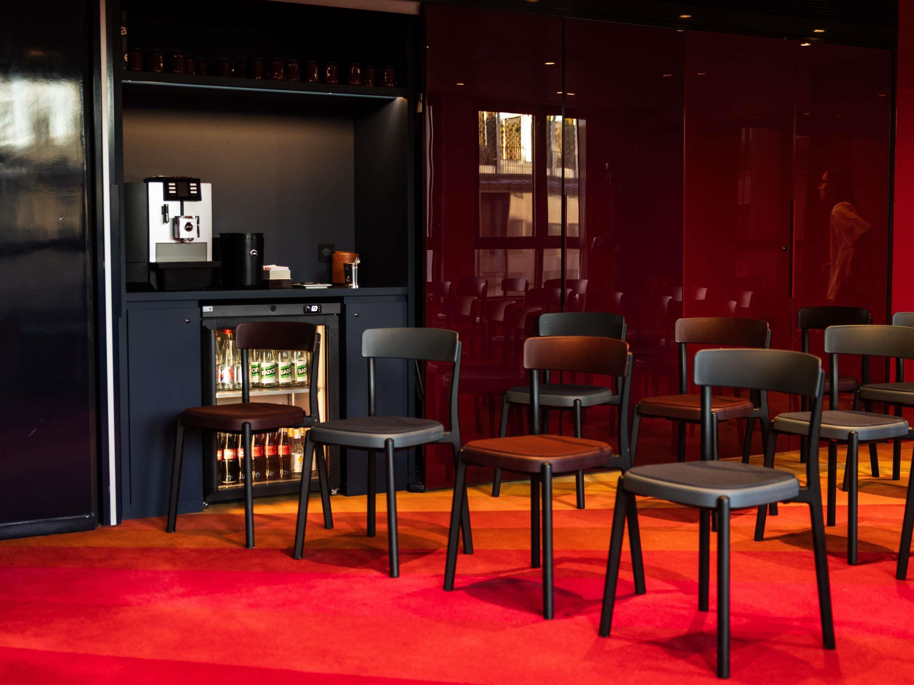 A bar area with a refrigerator stocked with bottles, a coffee machine, and a shelf with glasses. There are several chairs arranged for seating.