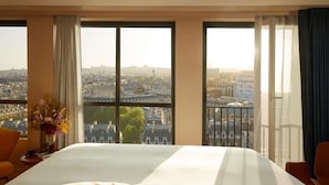 A hotel room with a large bed, two windows with curtains, a vase with flowers, and a view of a cityscape.