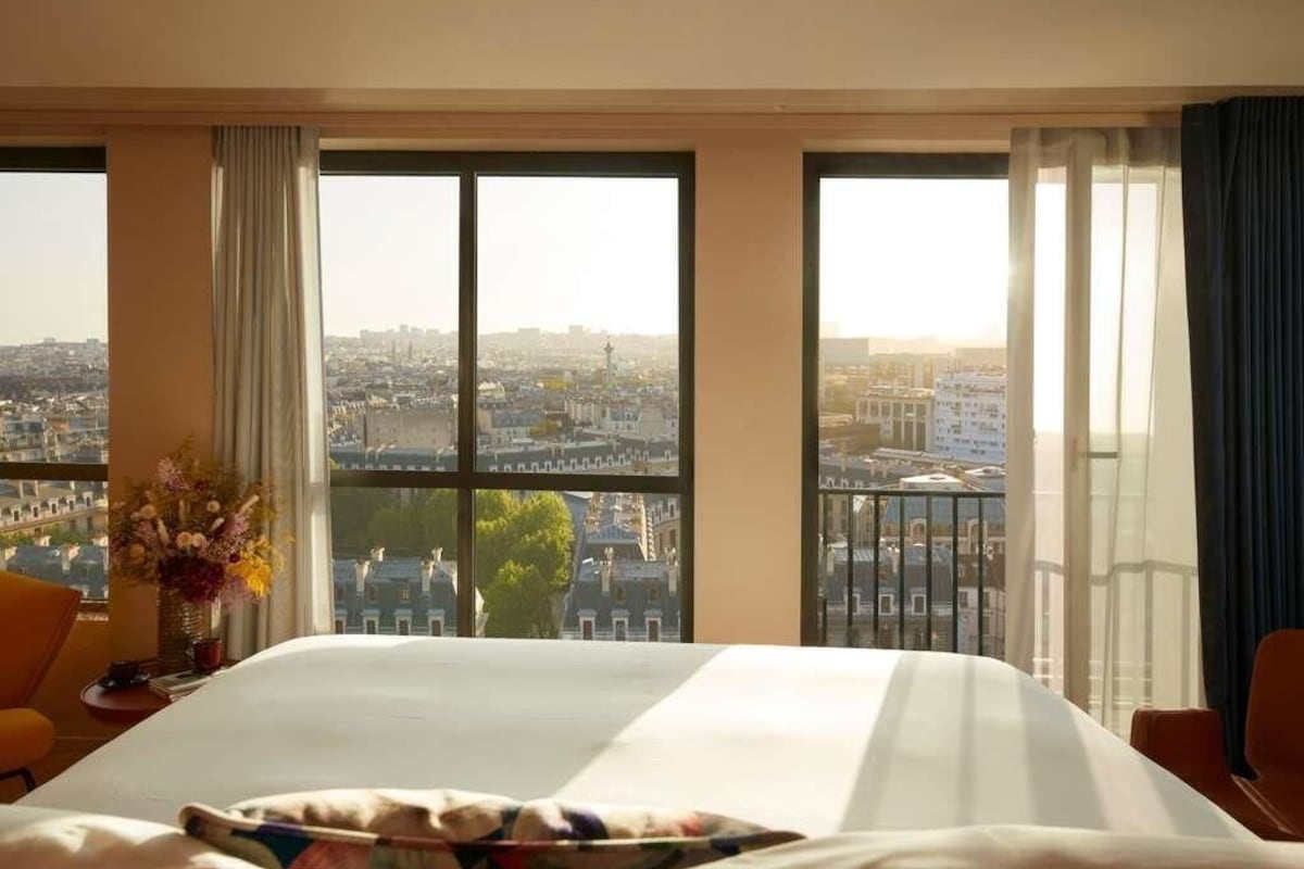 A hotel room with a large bed, two windows with curtains, a vase with flowers, and a view of a cityscape.