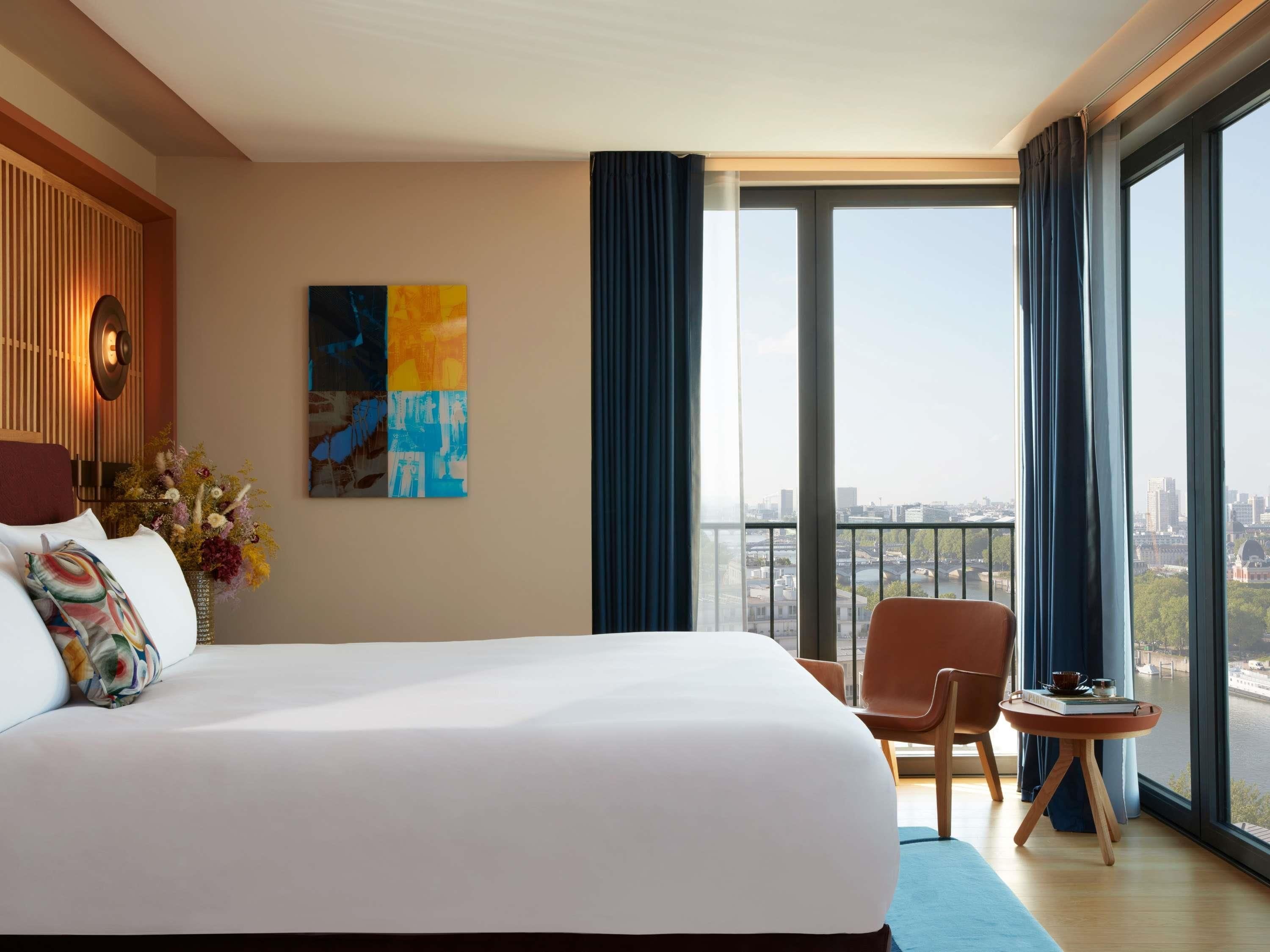 A hotel room with a large bed, a small table with a chair, a painting on the wall, and a view of a cityscape through large windows.