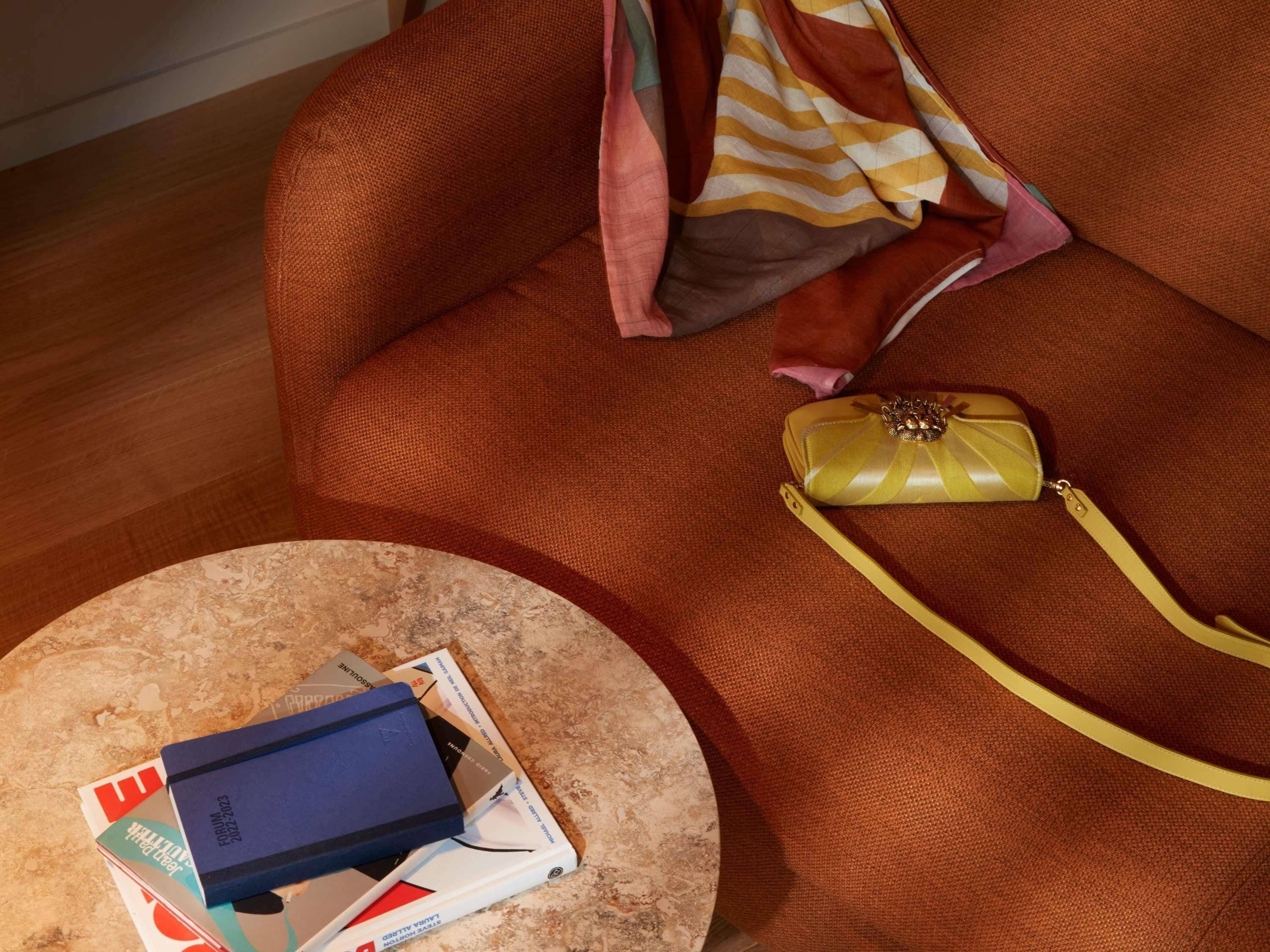 A round table with a stack of magazines and a blue book, a brown sofa with a yellow handbag, and a pink pillow.