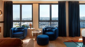 A modern living room with a city view, featuring two blue armchairs, a small round table with a book, and a brown sofa.