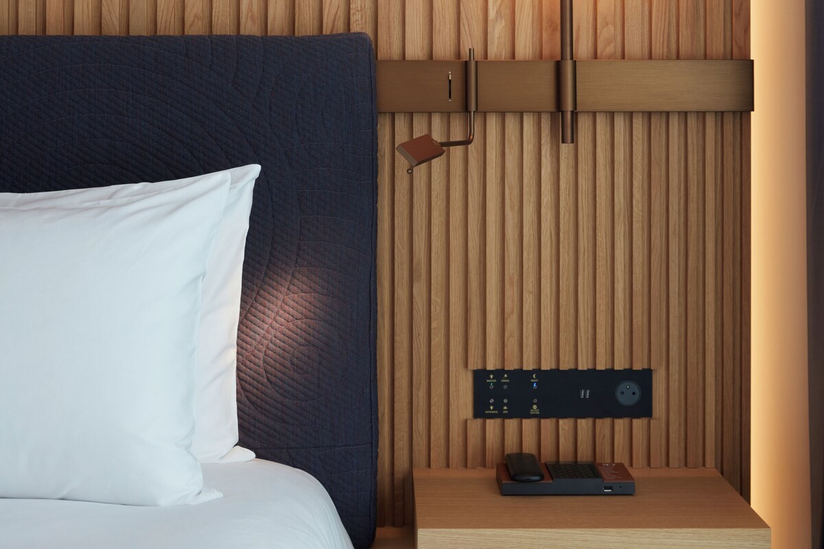 A modern hotel room with a wooden panel wall, a circular light fixture, a bedside table with a power strip, and a neatly made bed.