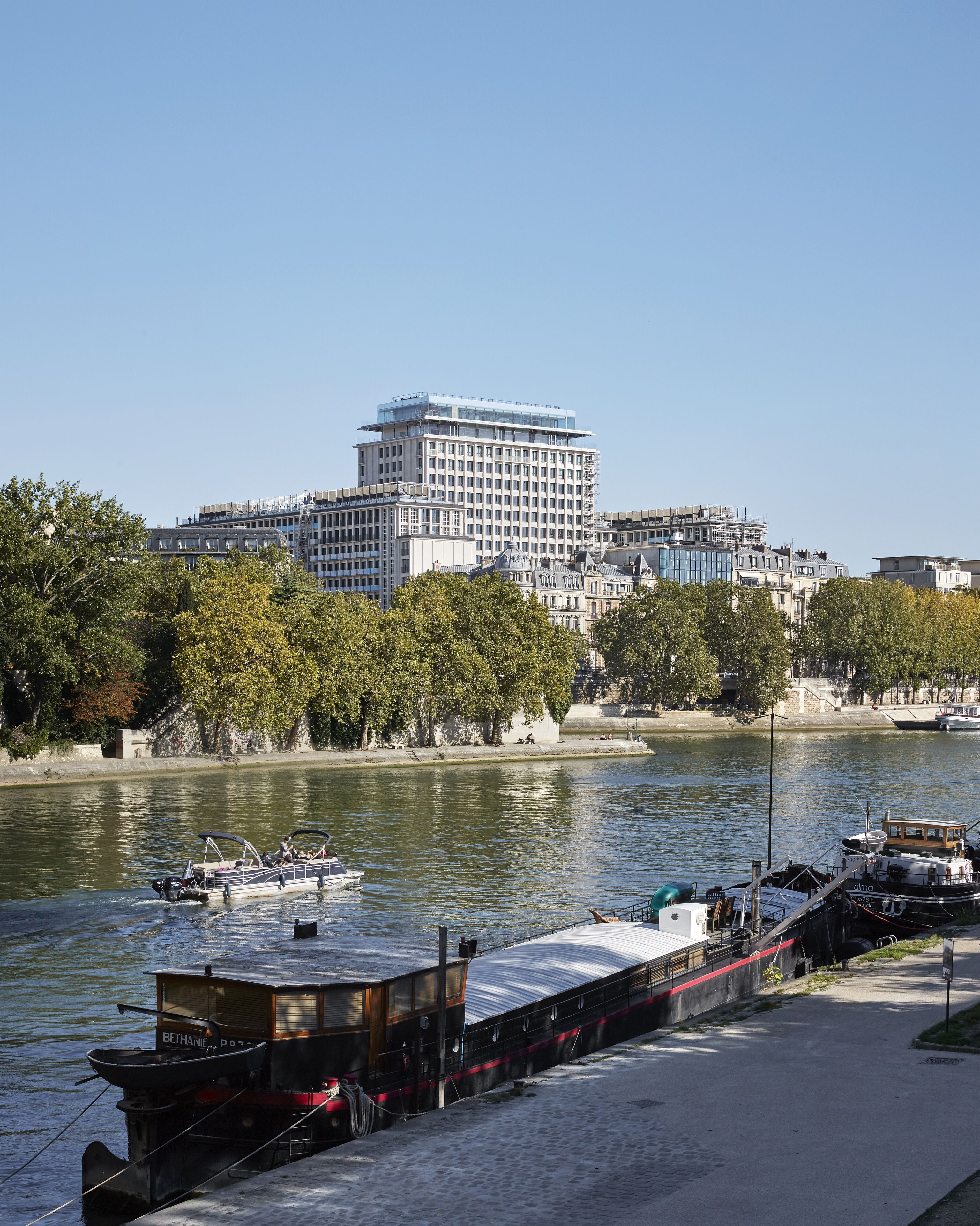 A river with boats docked along the shore, a large building in the background, and trees lining the riverbank.