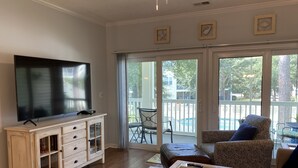 A living room with a flat-screen TV on a white cabinet, a patterned sofa, a coffee table with a bowl, and a sliding glass door leading to an outdoor area with a pool. - Ocean Keyes 3 BR Condo, XL balcony, pool, 1 block to beach, food,  & famous OD (North Myrtle Beach)