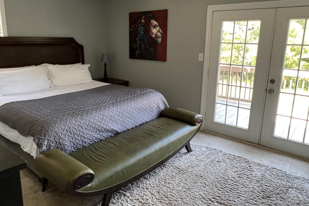 A bedroom with a bed, a green bench, a ceiling fan, a painting, and a door with a view.