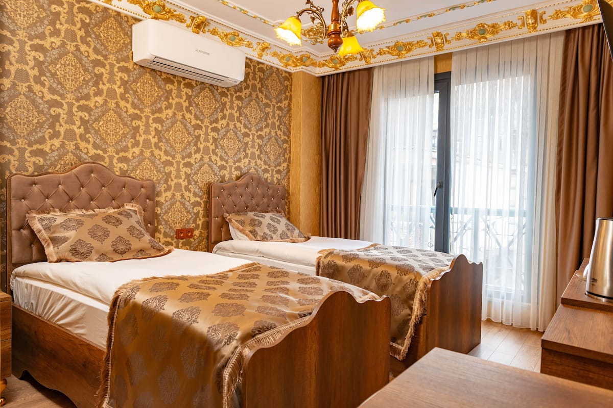 A hotel room with two beds, ornate wallpaper, a chandelier, and a view of the outdoors.