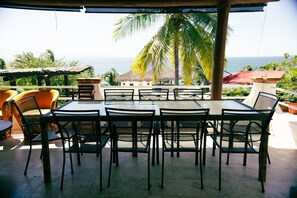 An outdoor dining area with a table and chairs, overlooking a scenic view of the ocean and palm trees. - Private Beach- Amazing Surf- Pool -Staff - Gated Home- Suburban & Chef Available (Punta Mita)
