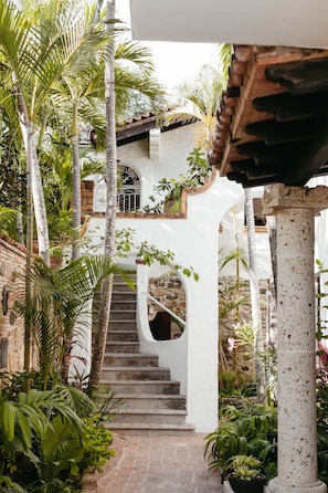 Eine Treppe, umgeben von üppiger Vegetation, führt zu einem weißen Gebäude mit steinernen Fundamenten und einem Metallgitter.