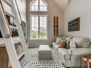 Un salón acogedor con un ventanal grande, un sofá gris con muchos cojines, una alfombra estampada y una escalera de madera.