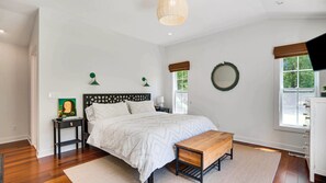 A bedroom with a bed, bedside tables, a wooden bench, a wall-mounted mirror, and a window with a view of greenery.