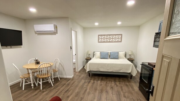 A compact room with a bed, a dining table with chairs, a red sofa, an air conditioning unit, and a TV mounted on the wall.